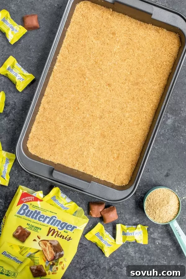 Preparing the graham cracker crust for Butterfinger S'mores Bars, showing crumbs being pressed into a baking pan