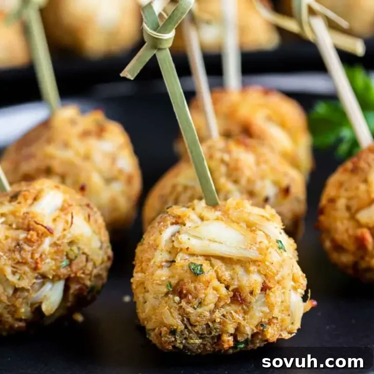 Close-up of golden-brown crab cakes on skewers, placed on a black plate, garnished with a sprig of parsley in the background.