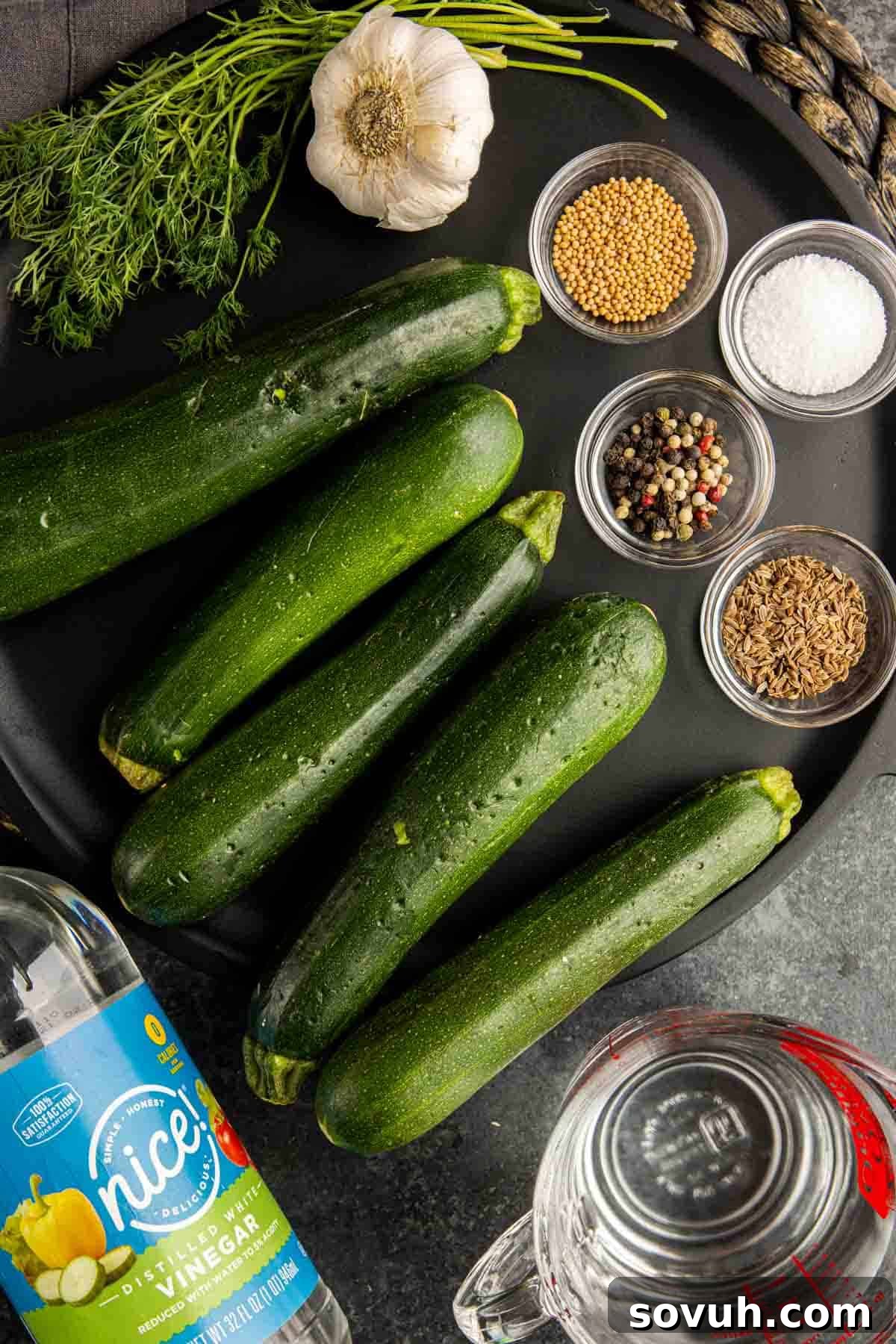 Quick Pickled Zucchini ingredients arranged on a dark surface, including fresh zucchini, dill, garlic, vinegar, and various pickling spices.