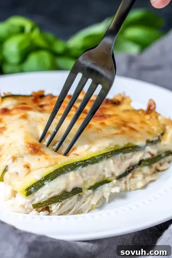 A fork taking a delicious, creamy bite out of a freshly baked Chicken Alfredo Zucchini Lasagna, showing the layers of chicken, zucchini, and cheese.