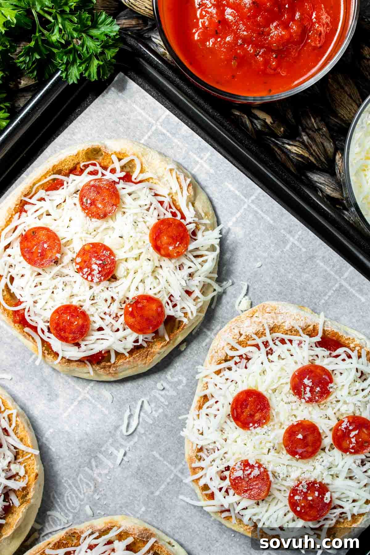 English muffin pizzas lined up on a baking sheet, topped with sauce, cheese, and pepperoni, ready for the oven.