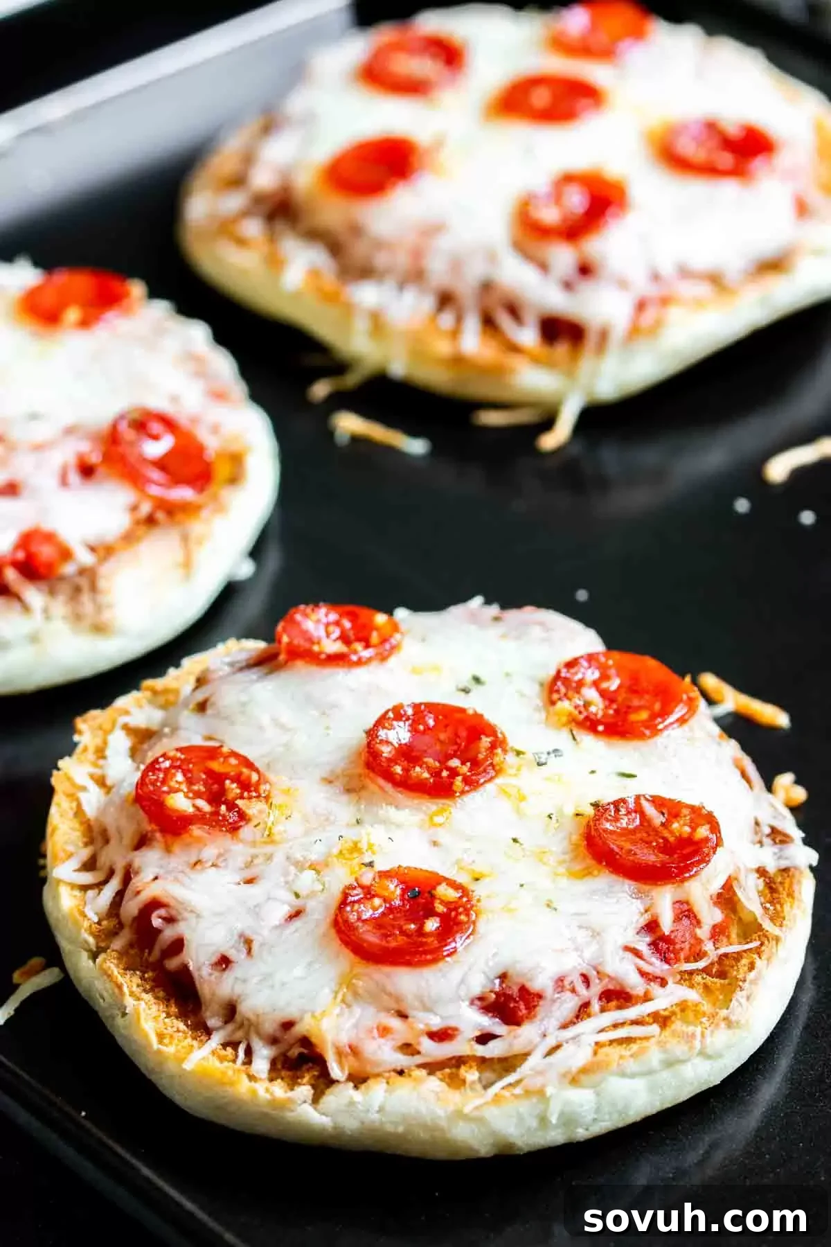 A baking sheet filled with English muffin pizzas, some with melted cheese and pepperoni, some plain.