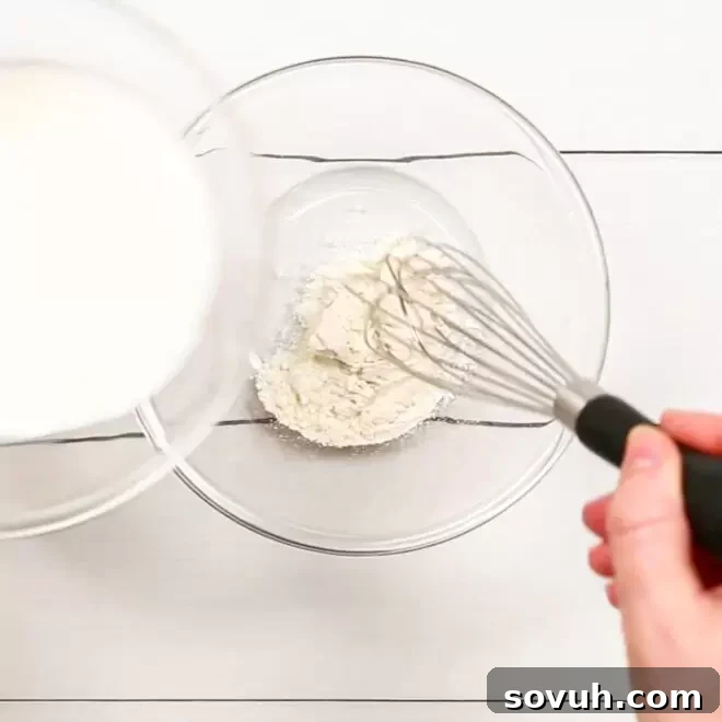 A person whisking flour and milk in a clear glass bowl, preparing the thickener for Creamed Corn.
