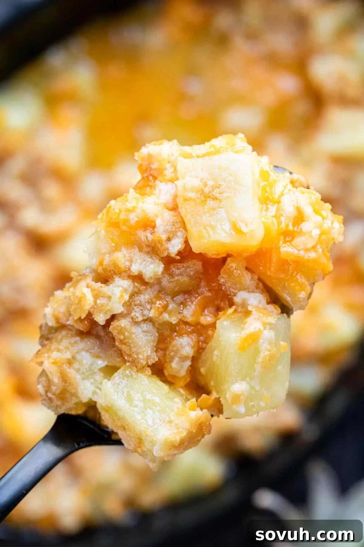 A scoop of golden pineapple casserole with a spoon, showing the cheesy, pineapple interior and crunchy cracker topping.