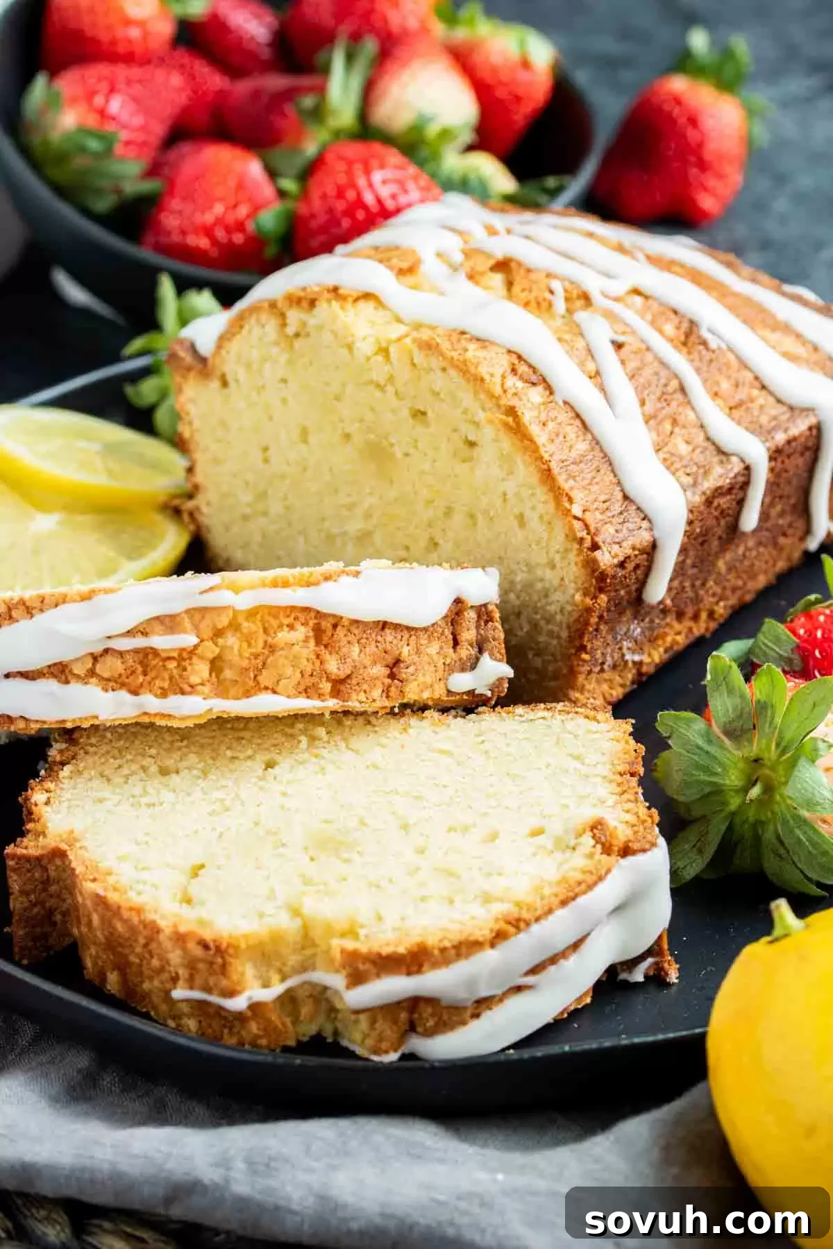 A whole lemon pound cake with elegant icing and fresh strawberries on a dark plate, inviting a taste.