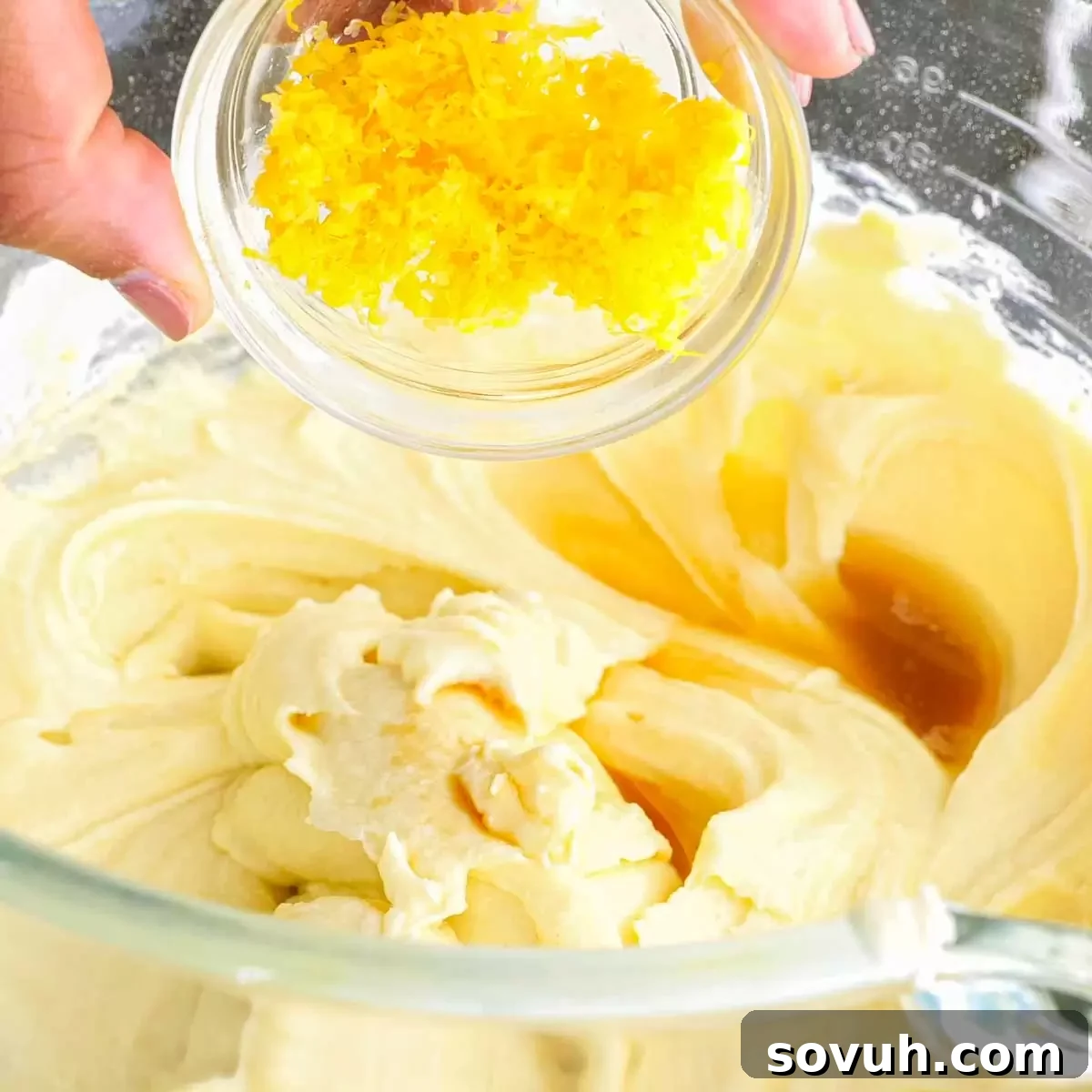 A hand adding fresh lemon zest to the cake batter in a mixing bowl, enhancing the citrus aroma.
