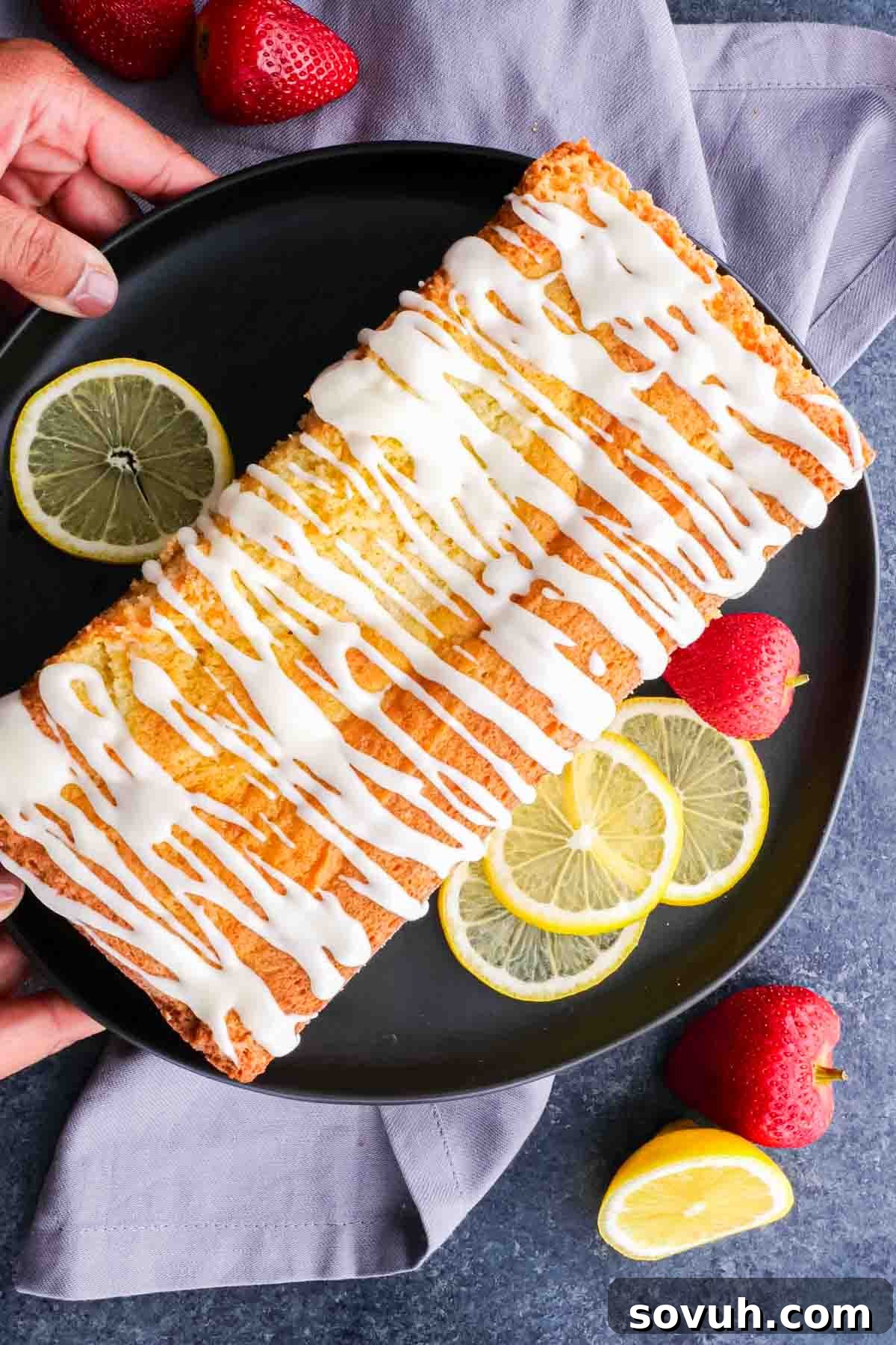 A perfectly sliced piece of lemon pound cake topped with lemon icing and fresh strawberries on a black plate.