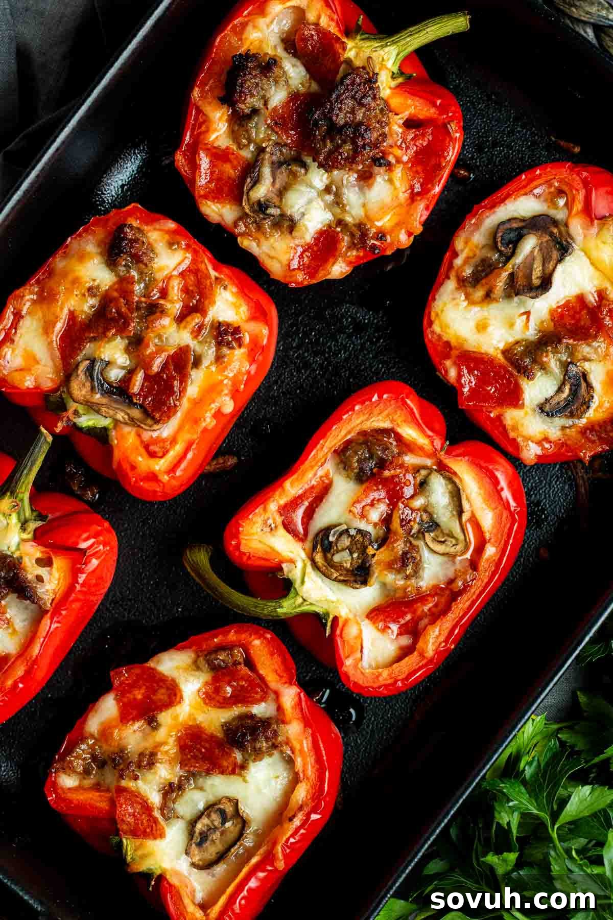 Red bell pepper halves filled with melted cheese, mushrooms, and browned meat, placed on a black tray, showcasing the delicious filling.