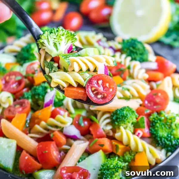 A close-up shot of a spoonful of colorful vegan pasta salad being lifted from a large bowl, showcasing fresh vegetables, pasta, and herbs.