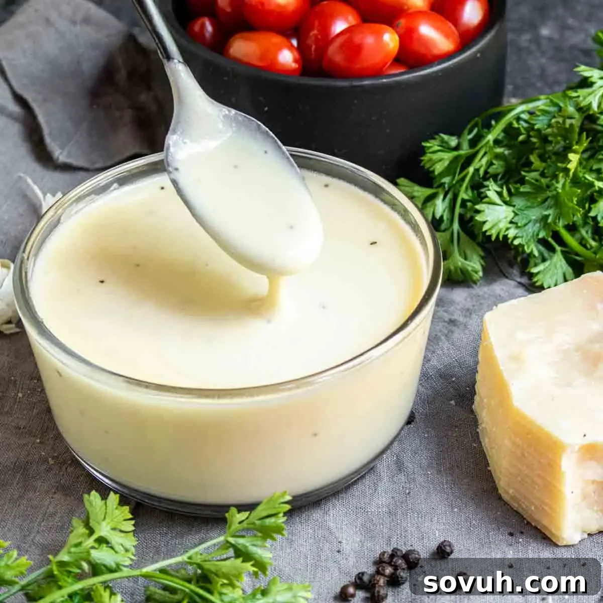 A bowl of rich and creamy Keto Alfredo Sauce, garnished with fresh parsley and a sprinkle of black pepper, on a rustic gray surface. Beside it are vibrant cherry tomatoes, a whole head of garlic, and a wedge of Parmesan cheese, highlighting the fresh ingredients.