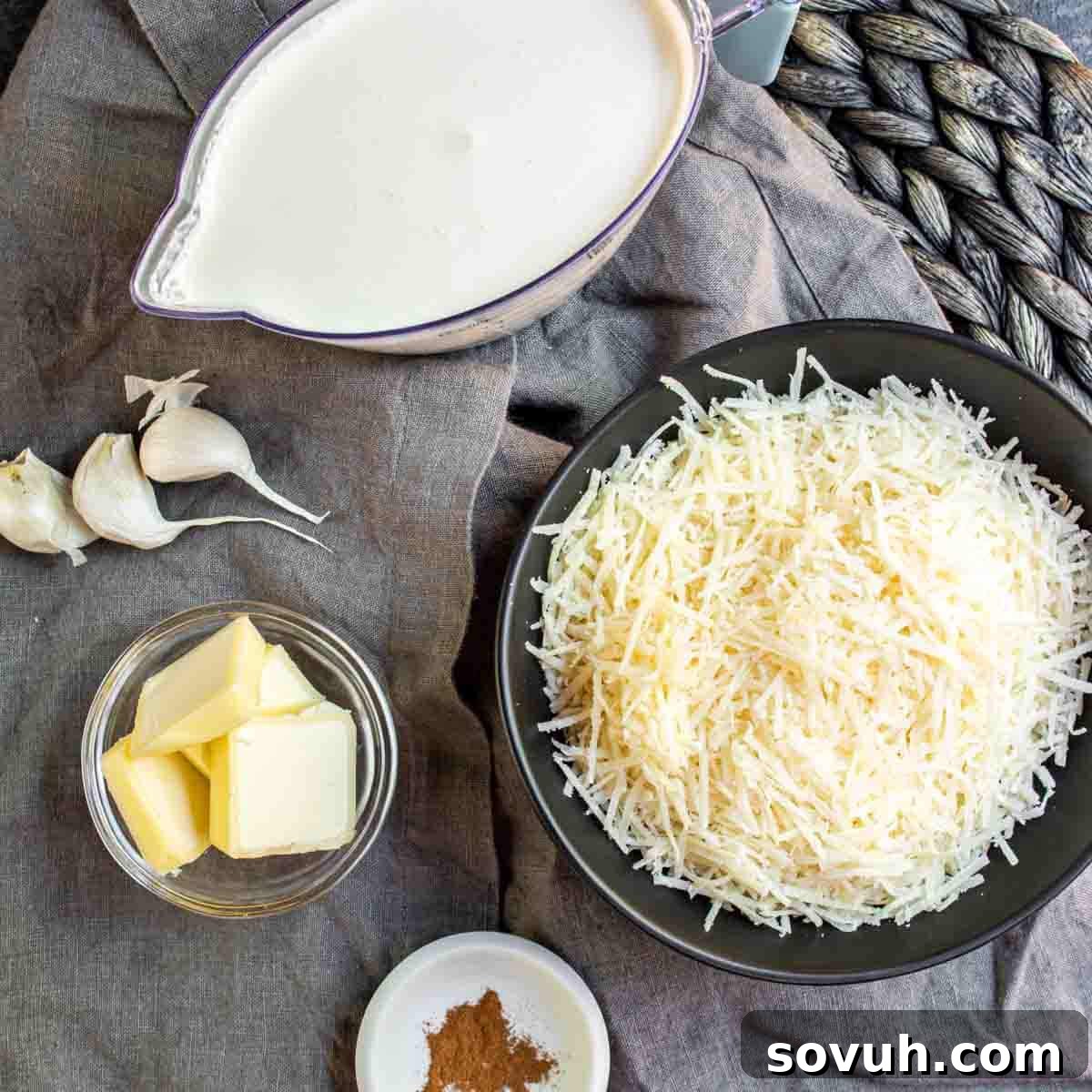 All the fresh ingredients laid out for making Keto Alfredo Sauce: a block of Parmesan cheese, a stick of butter, fresh garlic cloves, a small bowl of ground nutmeg, a jug of heavy cream, and salt and pepper shakers, all on a gray textured background.