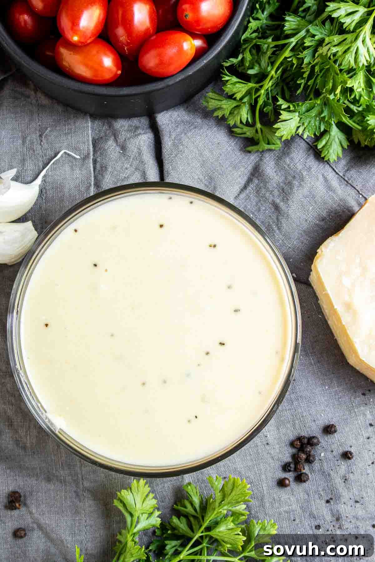 A beautifully plated bowl of creamy Keto Alfredo Sauce, ready to be served. It's surrounded by fresh ingredients like vibrant cherry tomatoes, fragrant parsley, a garlic bulb, and a generous wedge of Parmesan cheese, all on a textured gray background.