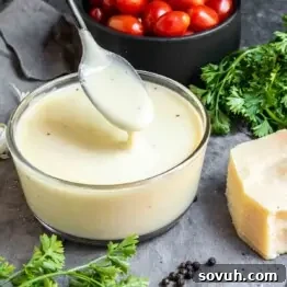 A bowl of Keto Alfredo Sauce with a spoon, alongside cherry tomatoes, parsley, and a wedge of parmesan cheese on a gray surface.