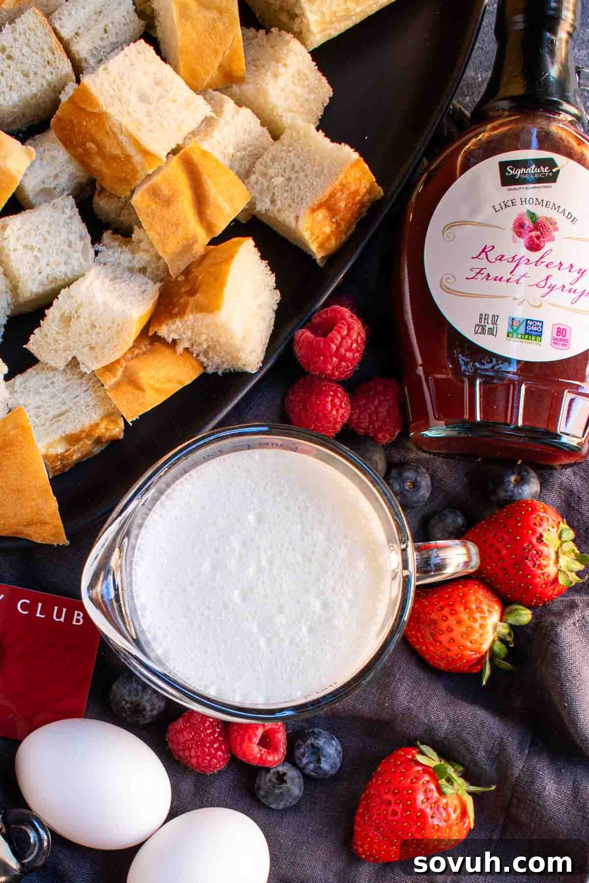 A bowl of maple syrup, a carton of eggs, and a small bowl of fresh mixed berries arranged on a black plate, ready to be used for Overnight French Toast Bake.