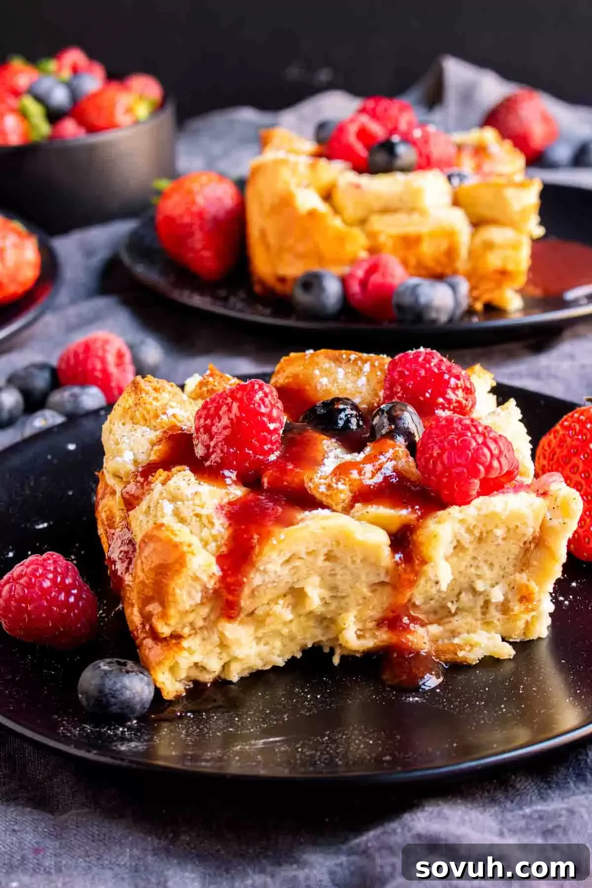 A perfectly portioned slice of Overnight French Toast Bake, generously adorned with fresh berries and a drizzle of syrup, resting on a black plate.