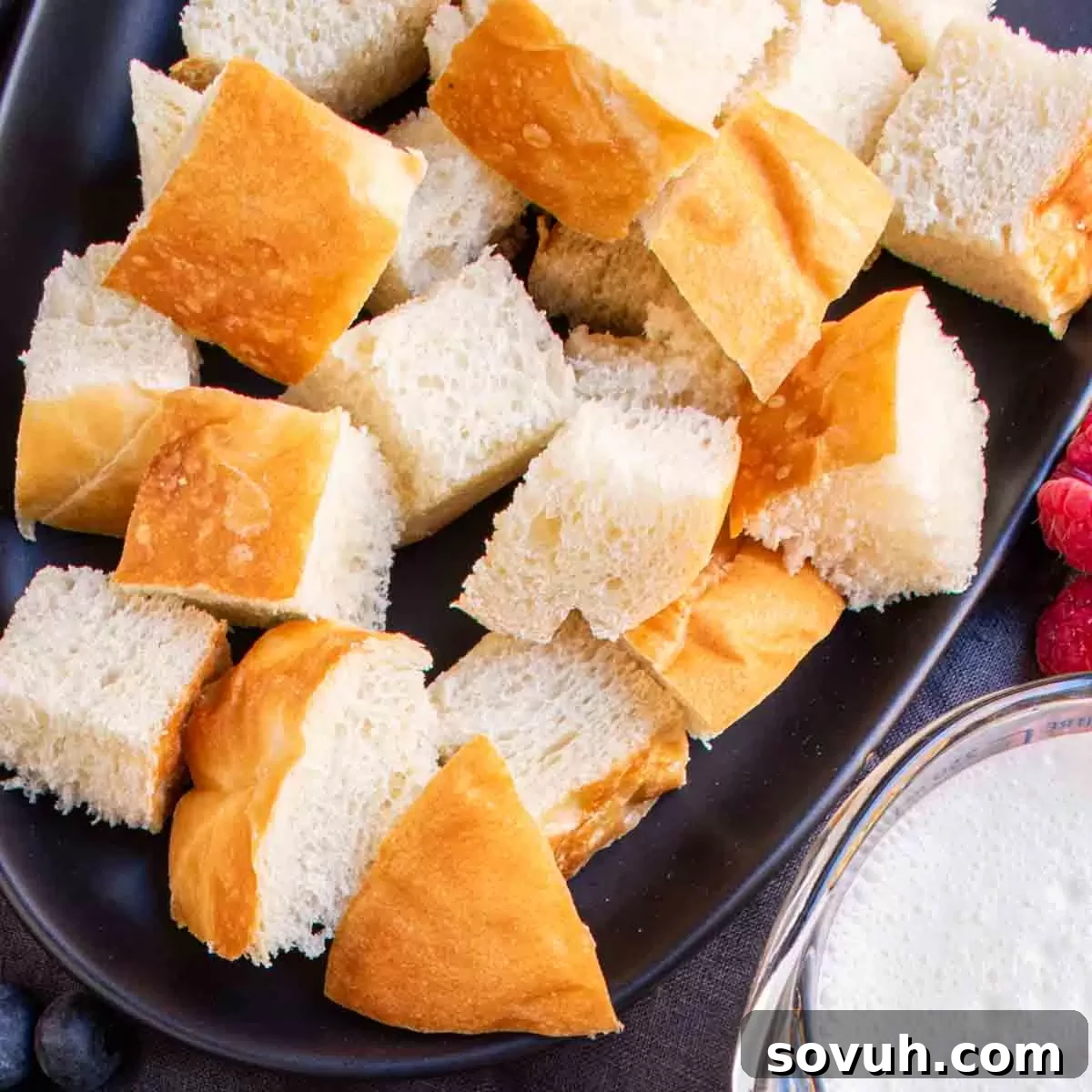 Bread cubes scattered on a black plate, accompanied by fresh berries and a pitcher of milk, representing the initial ingredients for Overnight French Toast Bake.