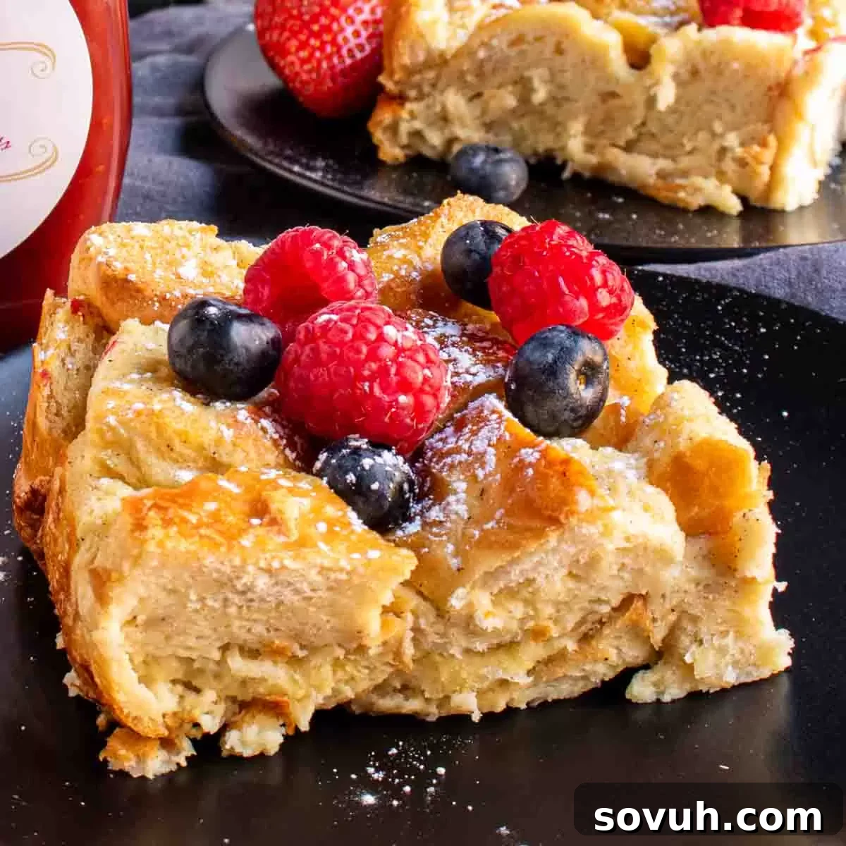 A close-up shot of a single slice of Overnight French Toast Bake, generously topped with an assortment of fresh berries and drizzled with sweet syrup, presented on a black plate.