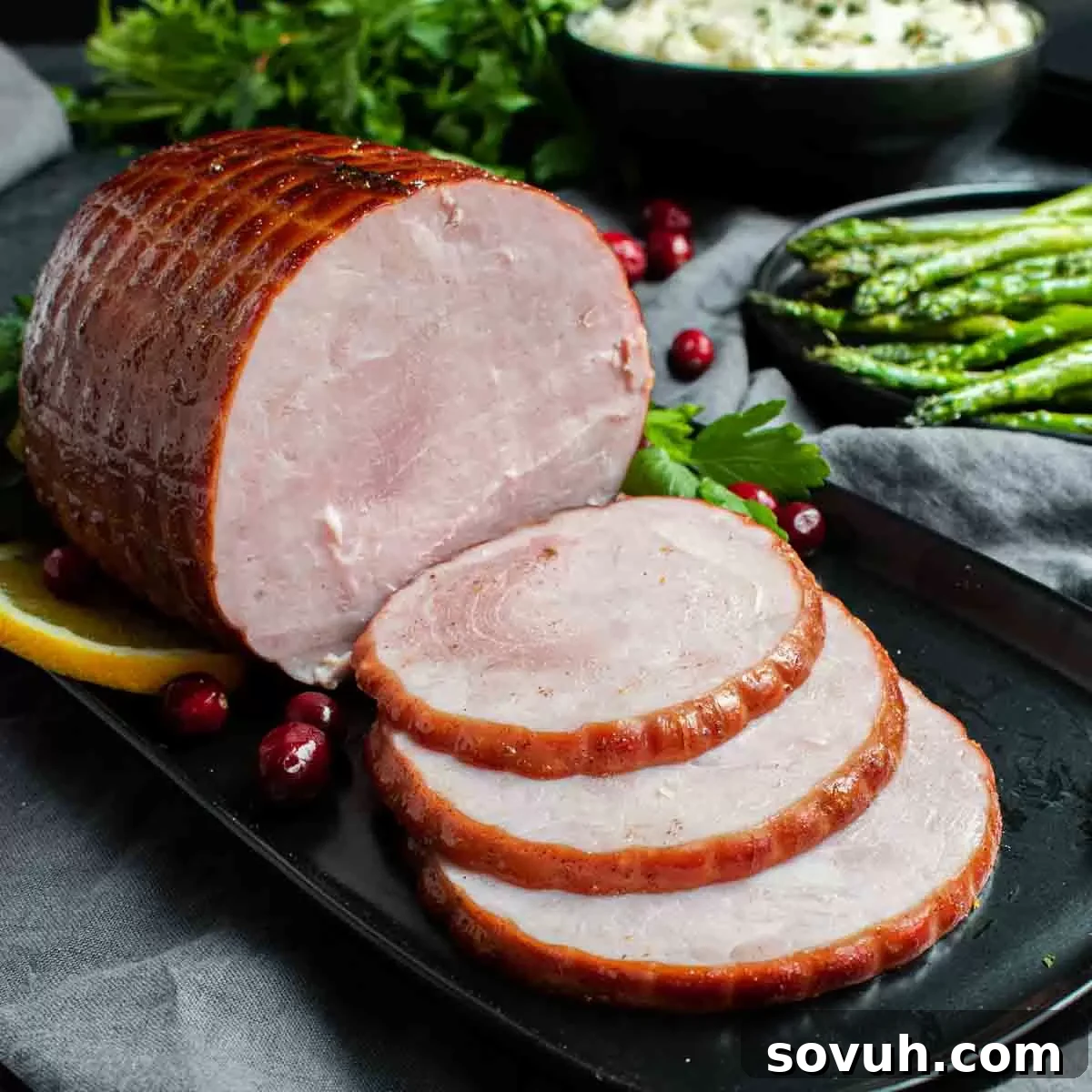 Sliced Instant Pot holiday ham on a black plate, garnished with fresh parsley and vibrant cranberries, with creamy mashed potatoes and crisp asparagus in the background.