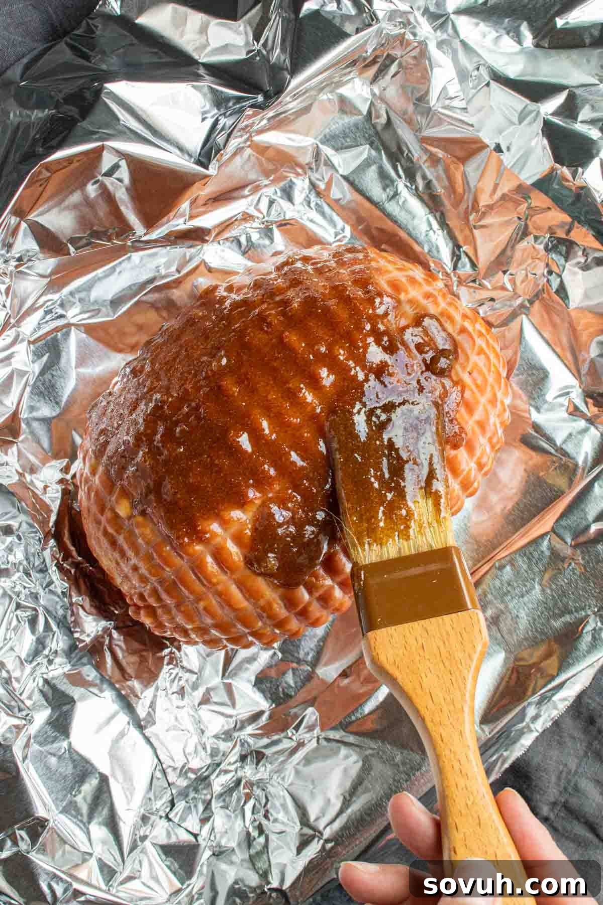 A hand expertly uses a basting brush to apply a glistening brown sugar glaze onto a scored ham, resting on a sheet of aluminum foil, ready for an easy Instant Pot holiday ham recipe.