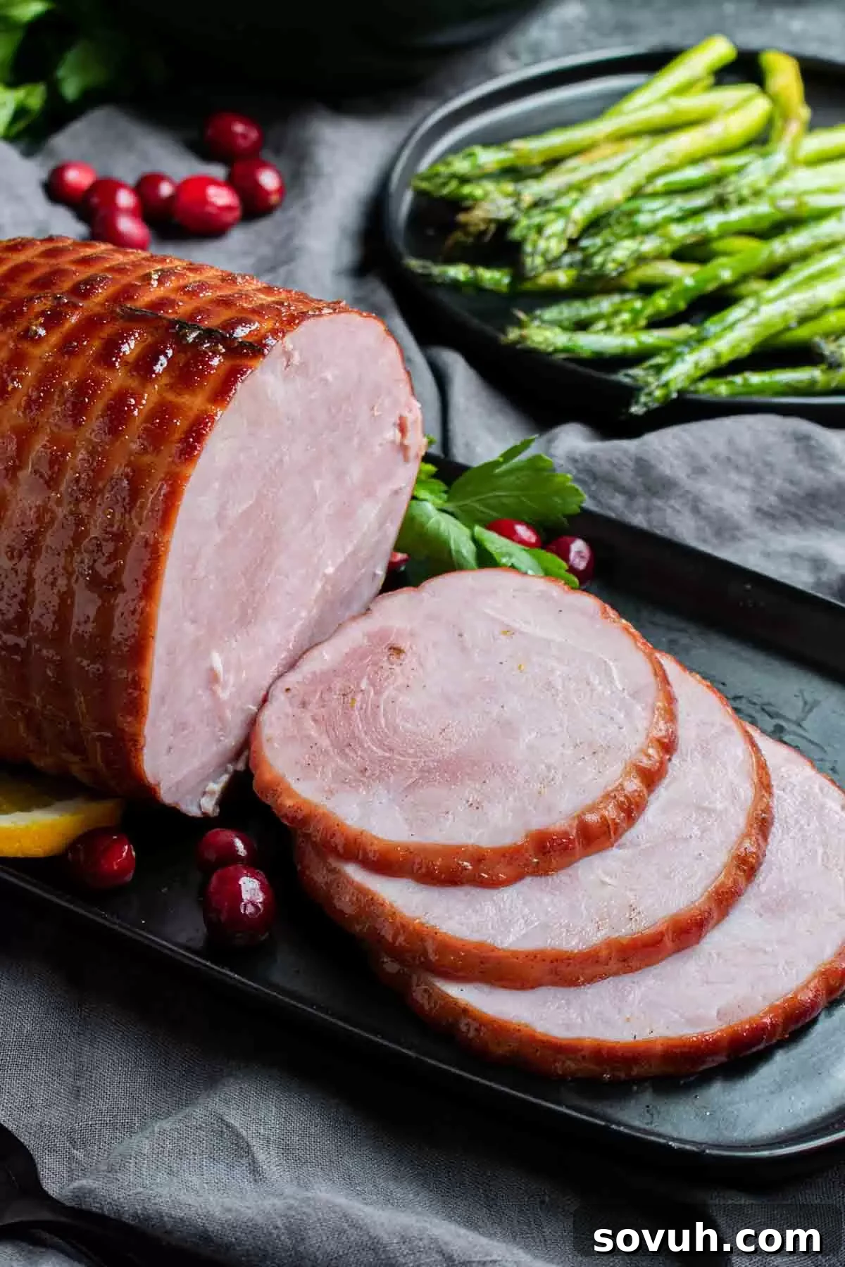 Perfectly sliced Instant Pot holiday ham on a black plate, artfully garnished with fresh cranberries and parsley, served alongside tender cooked asparagus.