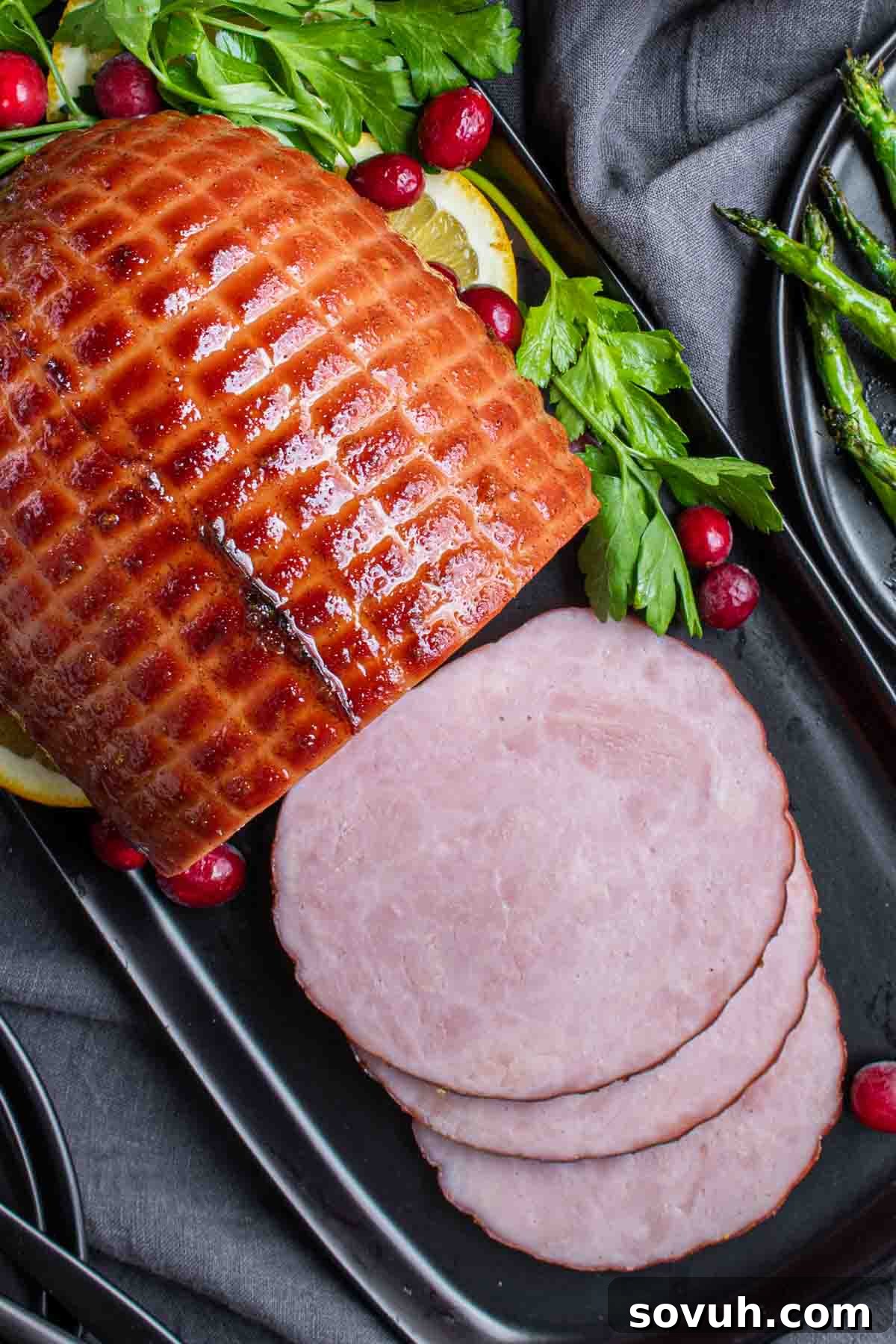 Beautifully sliced Instant Pot holiday ham presented on a black platter, garnished with fresh parsley, vibrant cranberries, and bright lemon slices, served alongside perfectly roasted asparagus.