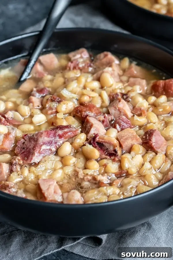 A hearty bowl of Navy Bean Soup, generously filled with chunks of pressure cooker ham and navy beans, with a spoon resting in the bowl.