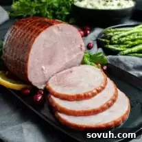 A cooked Easy ham recipe is partially sliced on a black plate, garnished with cranberries and lemon, with asparagus and mashed potatoes in the background.
