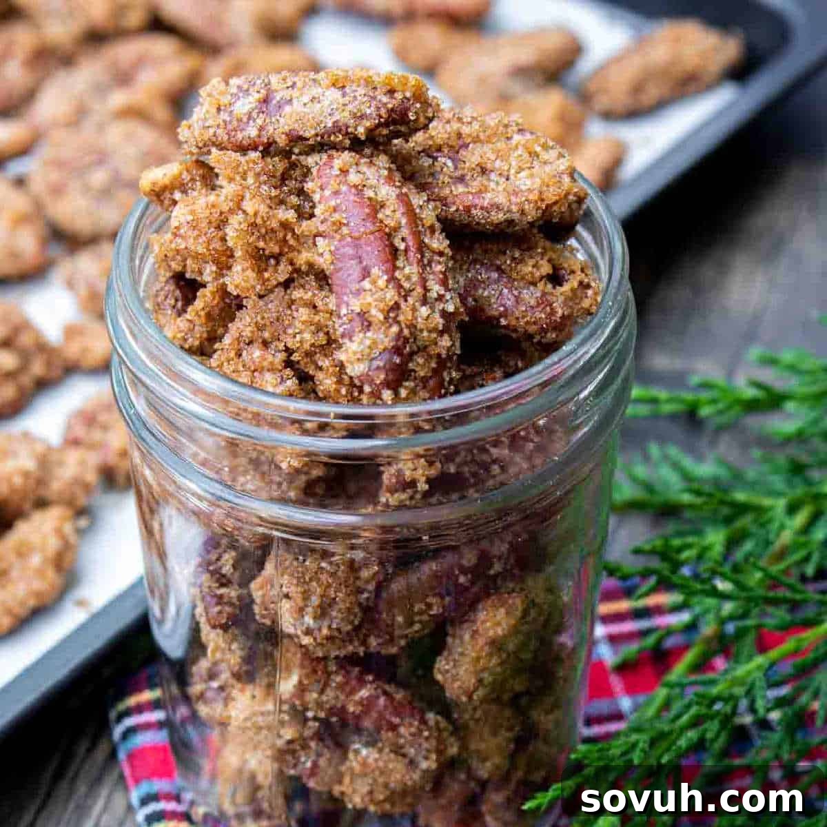 A glass jar filled with candied pecans sits on a tablecloth, with more pecans on a baking sheet in the background and sprigs of greenery nearby, highlighting a perfect homemade Christmas gift.