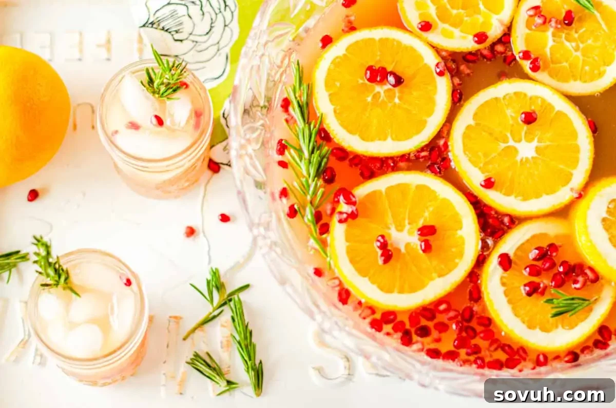A beautiful glass punch bowl filled with Christmas punch, garnished with vibrant citrus slices, ruby-red pomegranate seeds, and fresh green rosemary sprigs. Nearby, two small glasses, also garnished with ice and rosemary, await enjoyment, alongside whole, ripe oranges.