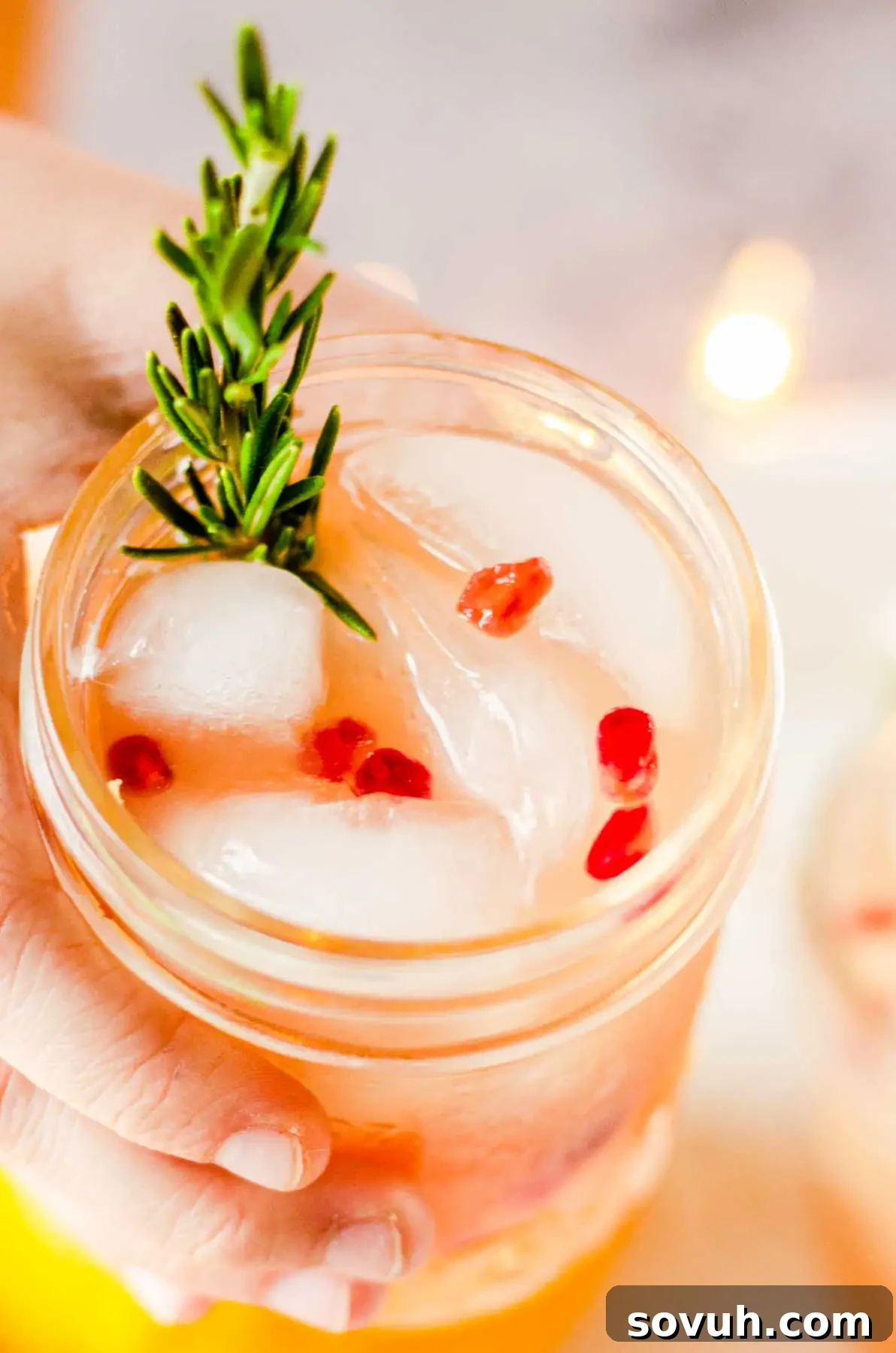 A hand holds a charming glass jar filled with the chilled Christmas punch, artfully garnished with glistening pomegranate seeds and a fresh sprig of rosemary, ready to be enjoyed.