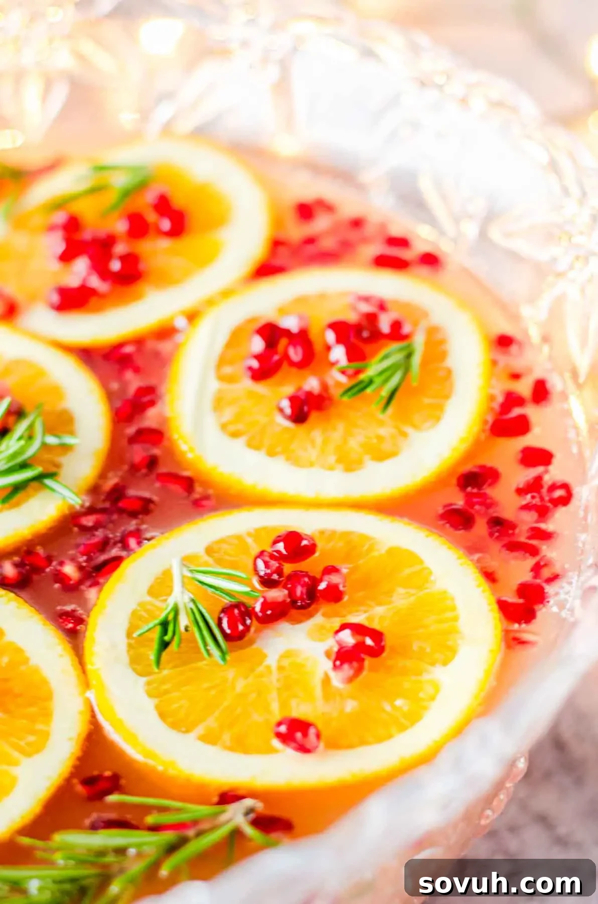 A close-up view of a vibrant punch bowl, showcasing a rich pinkish liquid adorned with floating orange slices, scattered pomegranate seeds, and fresh sprigs of rosemary, inviting a delightful taste.