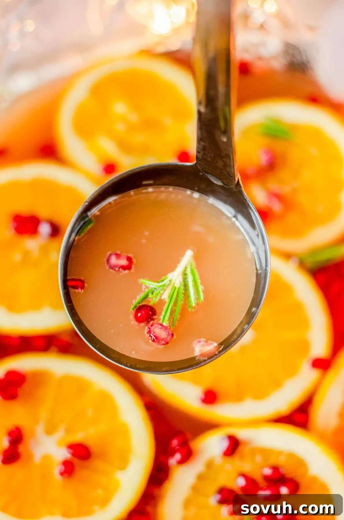 A ladle gracefully scoops out festive punch from a large glass bowl, revealing succulent orange slices, shimmering pomegranate seeds, and a fragrant sprig of rosemary suspended in the vibrant liquid.