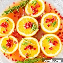A bowl of punch with orange slices, pomegranate seeds, and sprigs of rosemary floating on the surface.