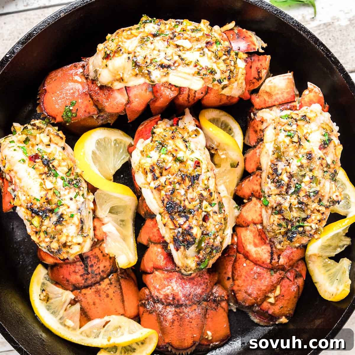 Top down photo of broiled lobster tails in a cast iron skillet, garnished with fresh herbs and lemon wedges