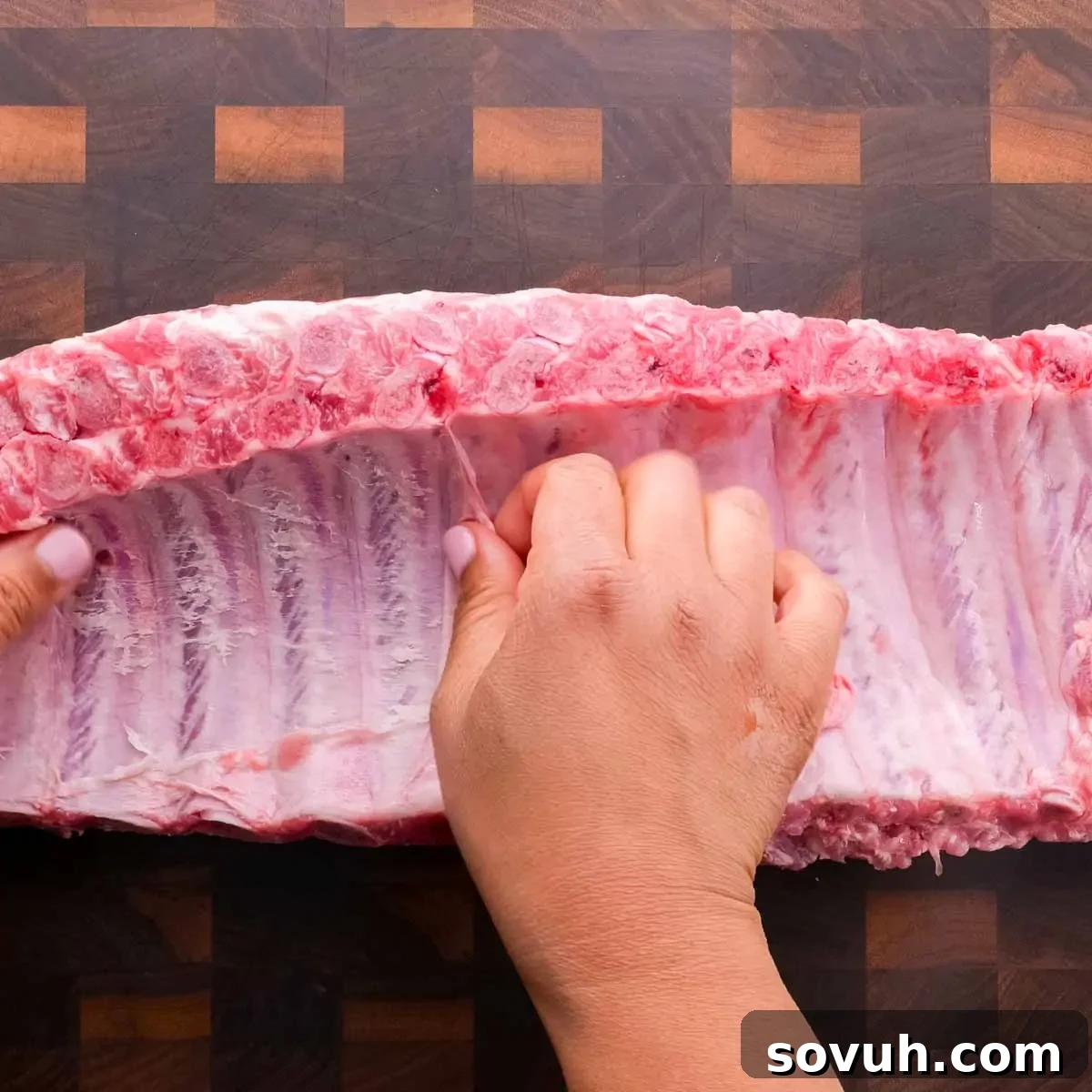 Hands trimming silk skin from a raw rack of St Louis-Style Ribs on a wooden cutting board.