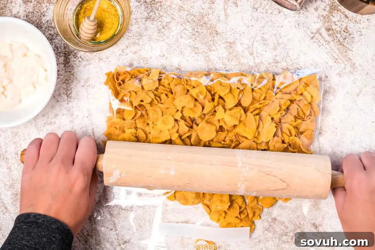 Person using a rolling pin to crush cornflakes in a sealed plastic bag on a floured surface, near a bowl of honey and a bowl of sauce.