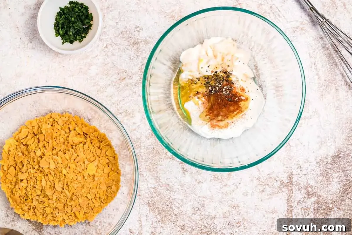 Two glass bowls on a countertop: one with crushed cornflakes, the other with a mixture of mayonnaise, spices, and herbs. Small bowl of chopped herbs and a whisk nearby.