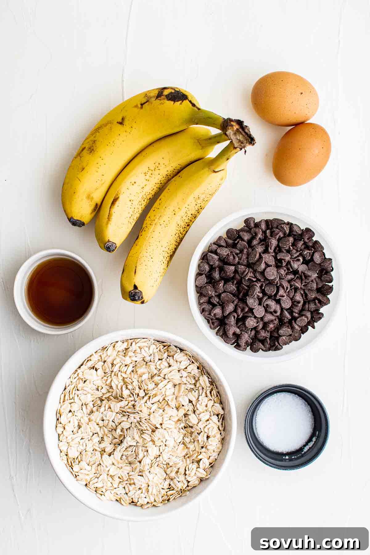 Ingredients laid out on a table: three ripe bananas, two eggs, a bowl of chocolate chips, a bowl of old-fashioned oats, a small bowl of salt, and a small bowl of vanilla extract.