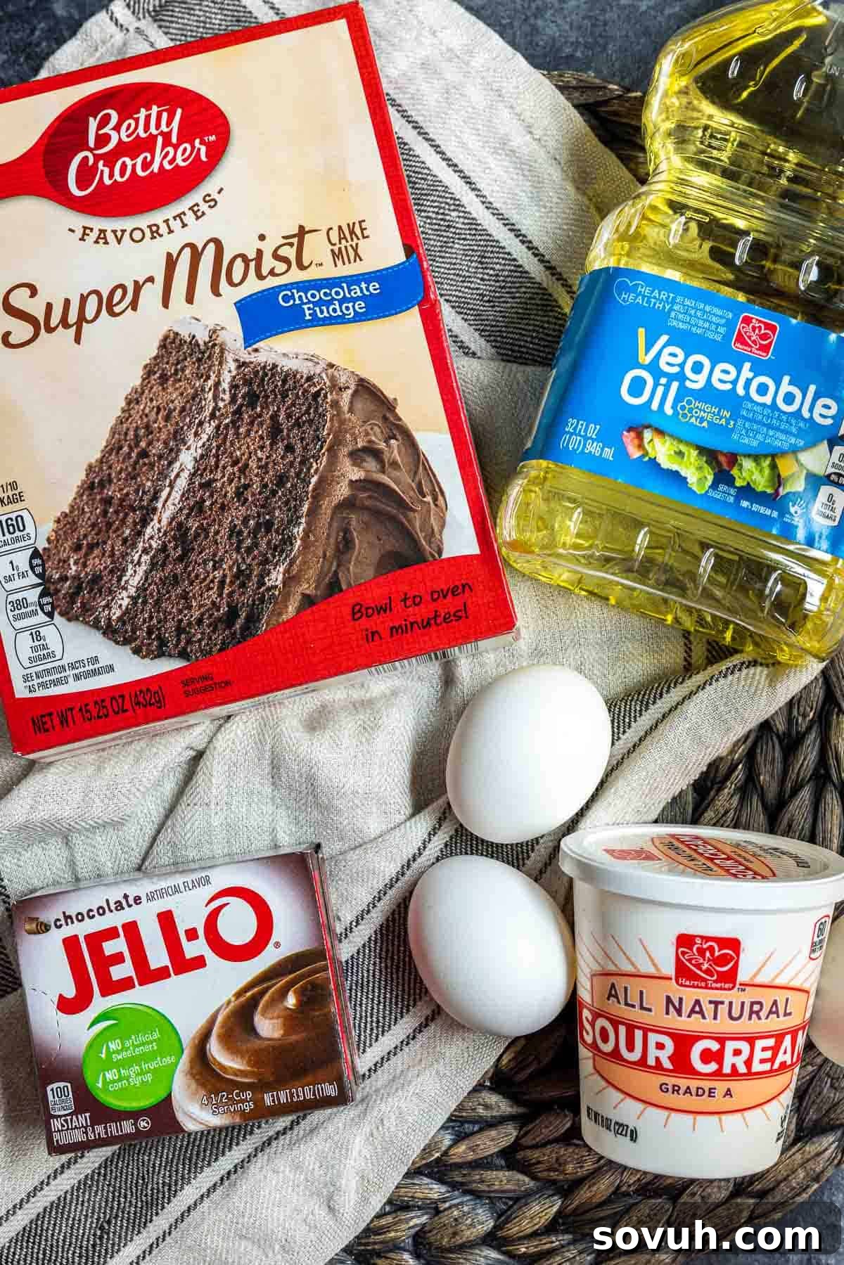 All the ingredients needed to bake a chocolate fudge bundt cake laid out on a table, including cake mix, pudding mix, sour cream, oil, eggs, chocolate, and heavy cream.