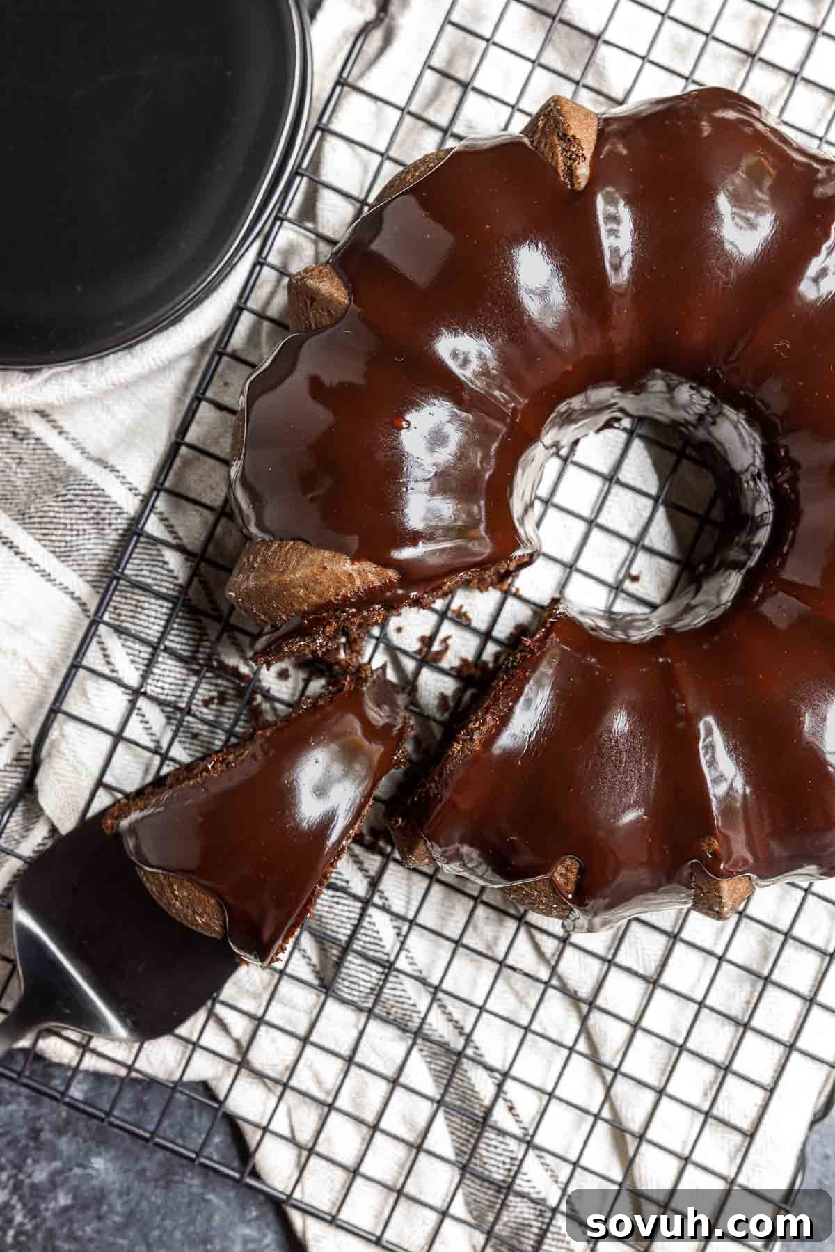 A slice of chocolate fudge cake with ganache glaze, served on a white plate with a fork.