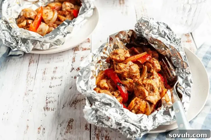 Two servings of cooked chicken with vegetables in aluminum foil packets are placed on white plates on a wooden table.