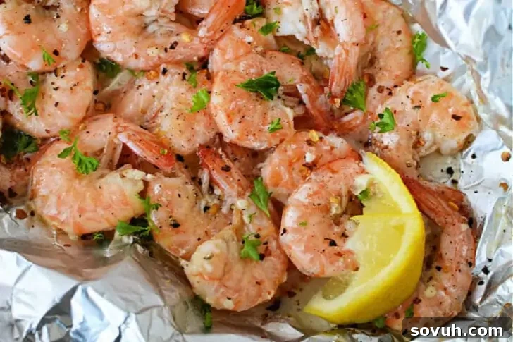 A close-up of seasoned, cooked shrimp garnished with parsley and served with lemon wedges on a foil surface.