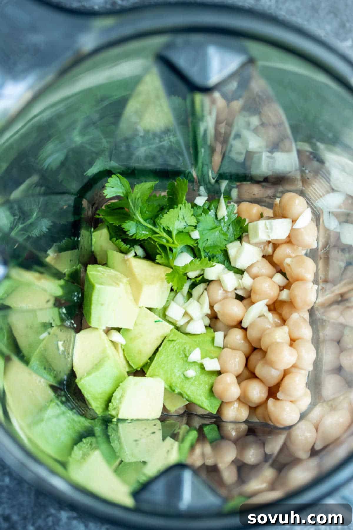 Fresh ingredients for Avocado Hummus, including chickpeas, avocado, cilantro, garlic, and lime, neatly arranged beside a blender.