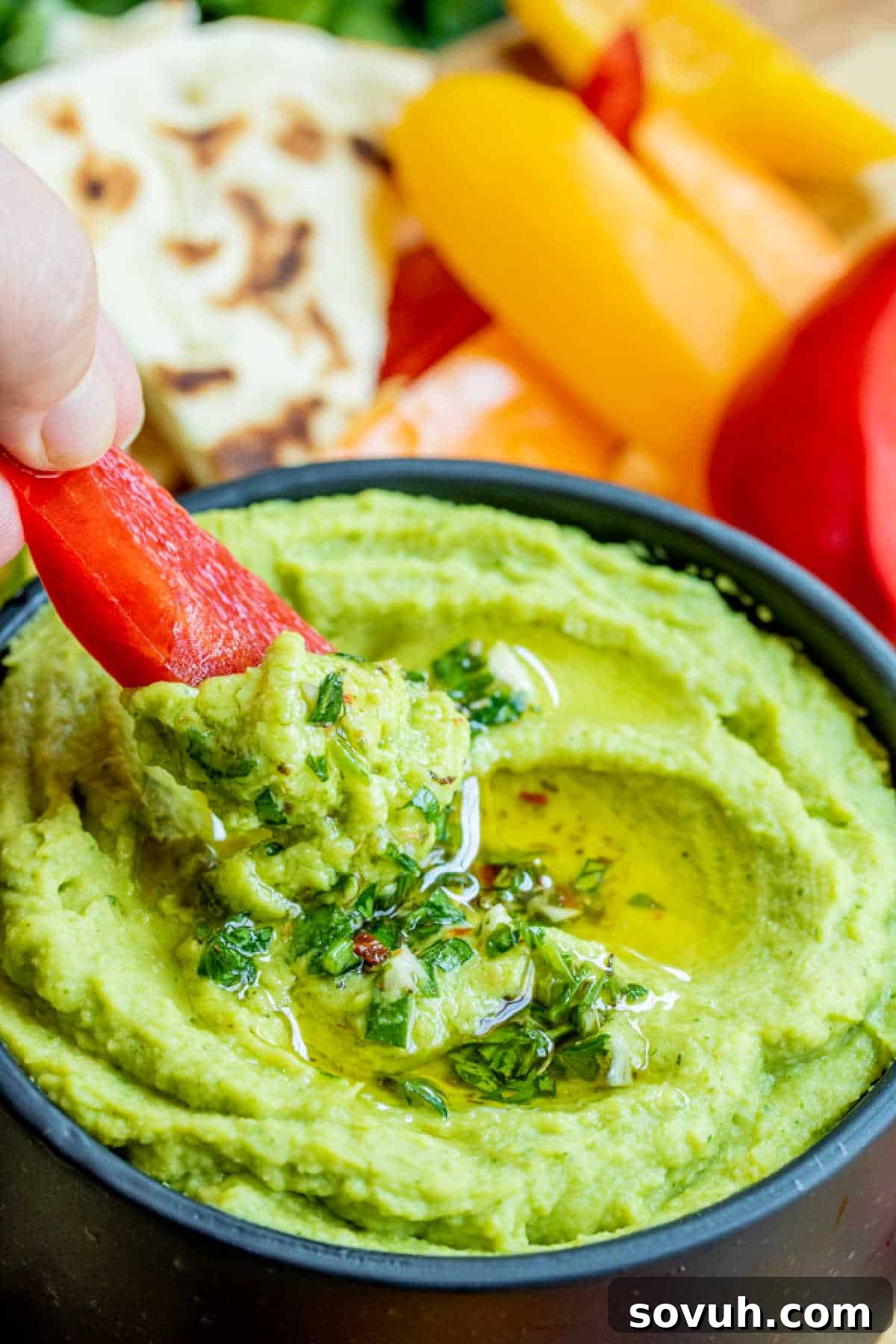 A vibrant red bell pepper slice being dipped into a bowl of creamy green Avocado Hummus.