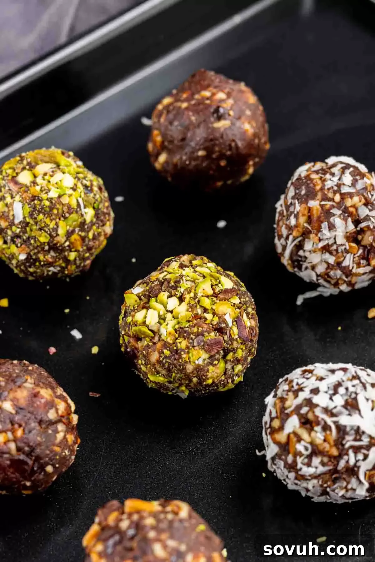 Bliss Balls 7 A close-up view of vibrant chocolate date balls rolled in finely chopped pistachios, neatly arranged on a light-colored baking sheet, showcasing their appealing green coating and uniform shape.