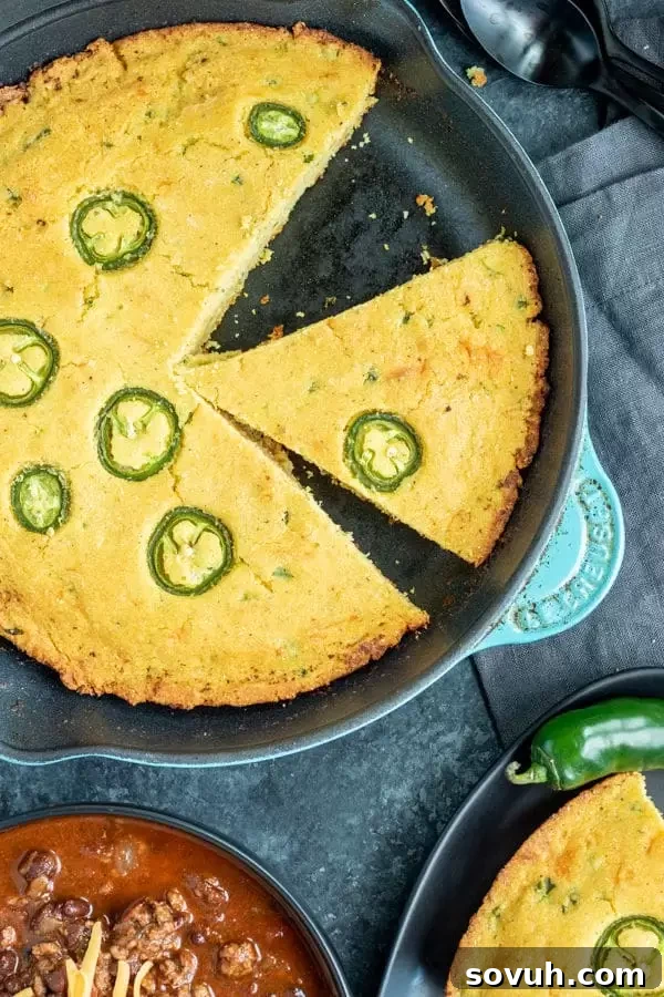 A cast iron skillet filled with freshly baked Jalapeno Cheddar Cornbread, ready to be sliced and served alongside a bowl of hearty chili.