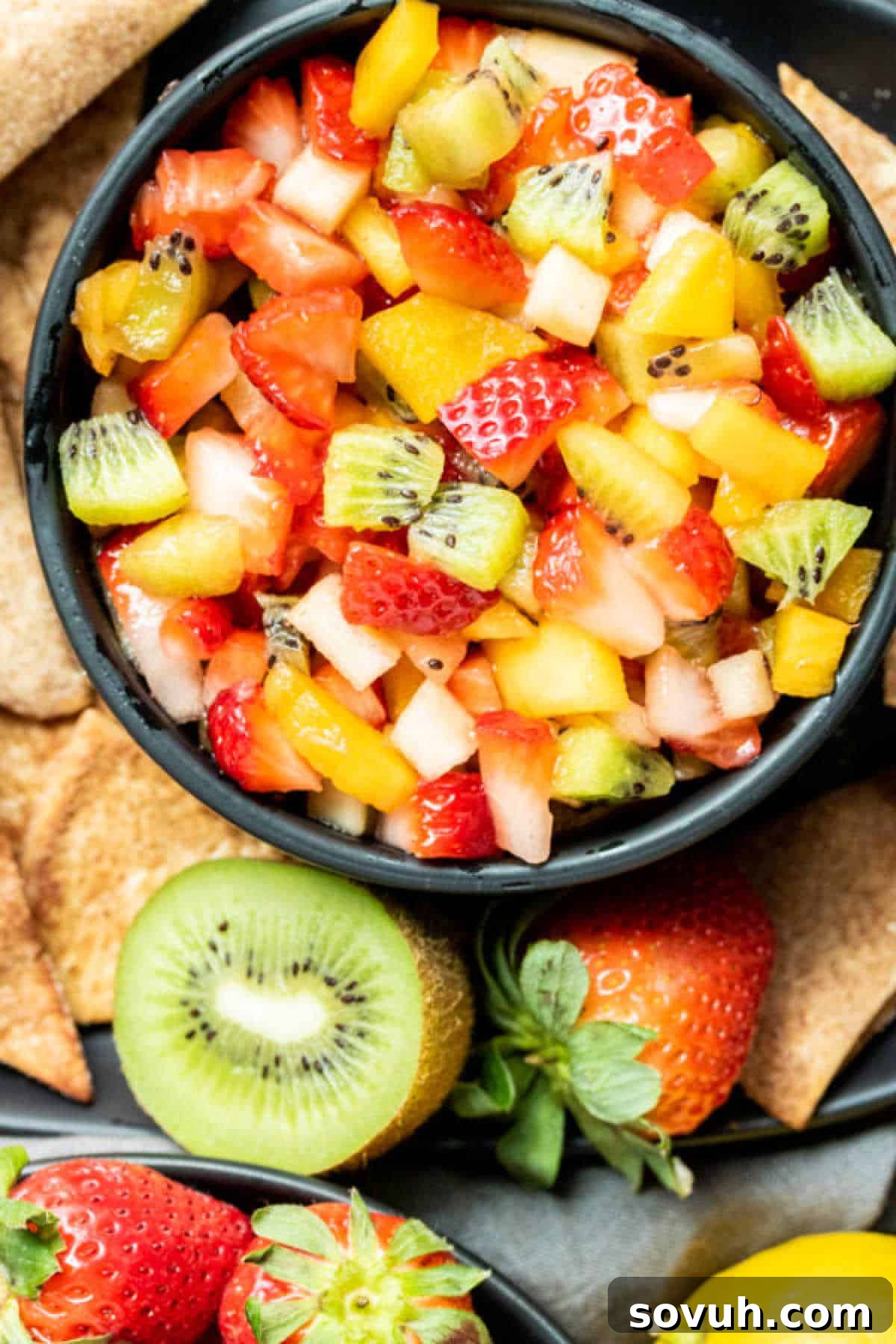 Top-down shot of fruit salsa in a black bowl surrounded by whole fruits