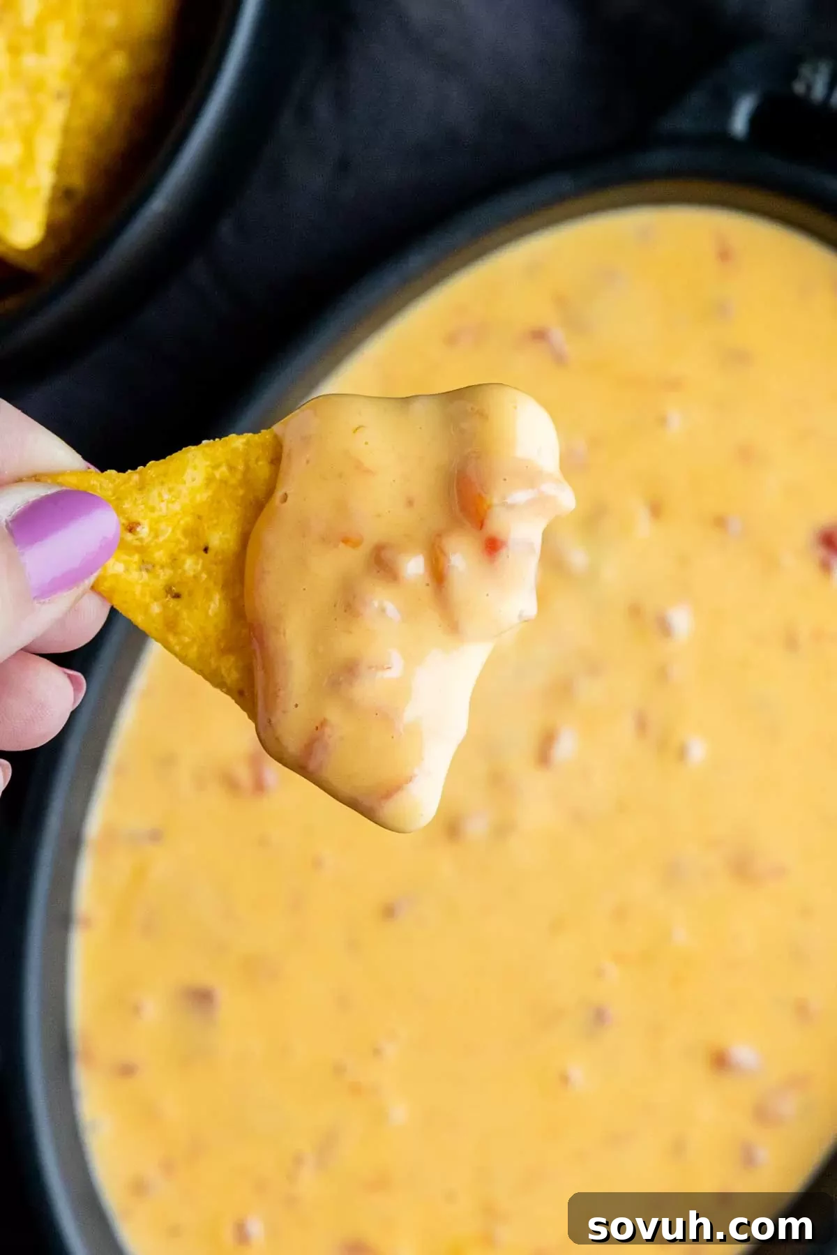 A hand holding a tortilla chip, generously scooped with warm, cheesy Rotel Dip, ready to be eaten.
