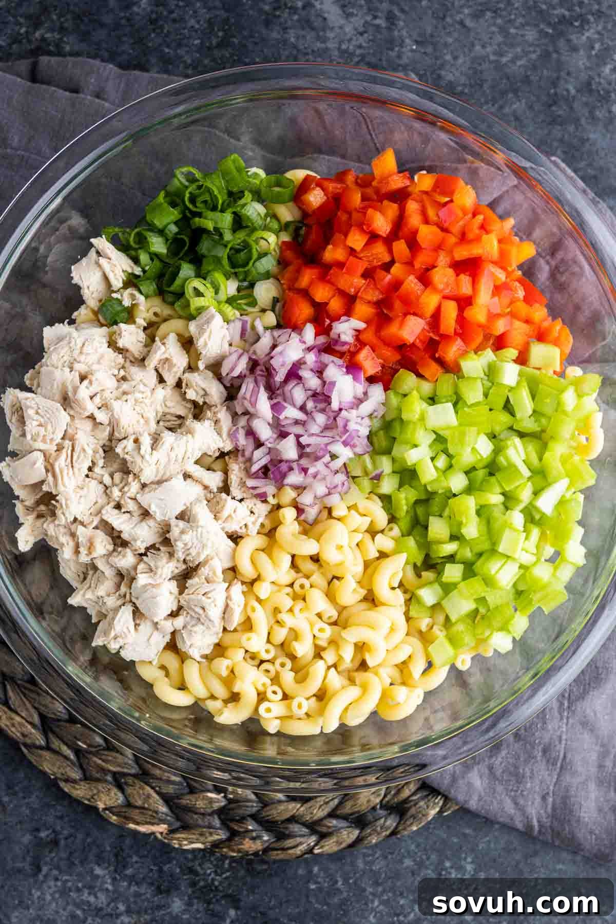 A large glass bowl filled with cooked elbow macaroni, chopped chicken, green onion, red bell pepper, celery, and red onion, before dressing is added.
