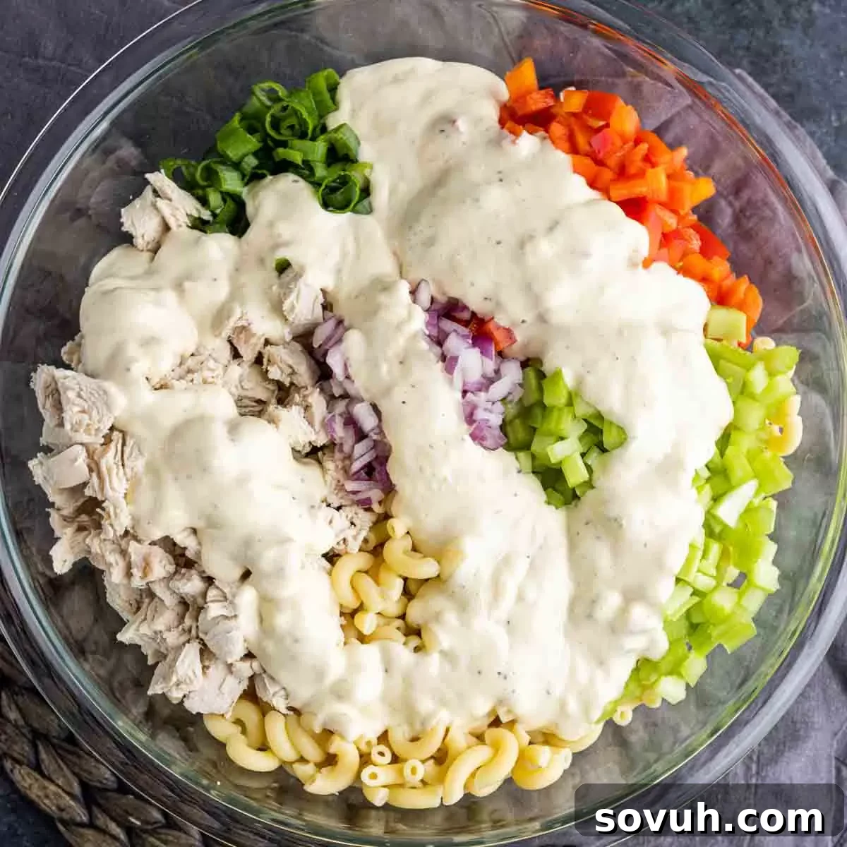 A large glass bowl with cooked macaroni, chicken, and chopped vegetables, with creamy dressing poured over and ready to be tossed.